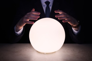 Photo of Businessman using glowing crystal ball to predict future at table in darkness, closeup. Fortune telling