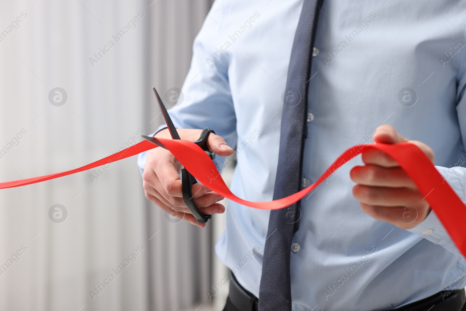 Photo of Man cutting red ribbon with scissors indoors, closeup