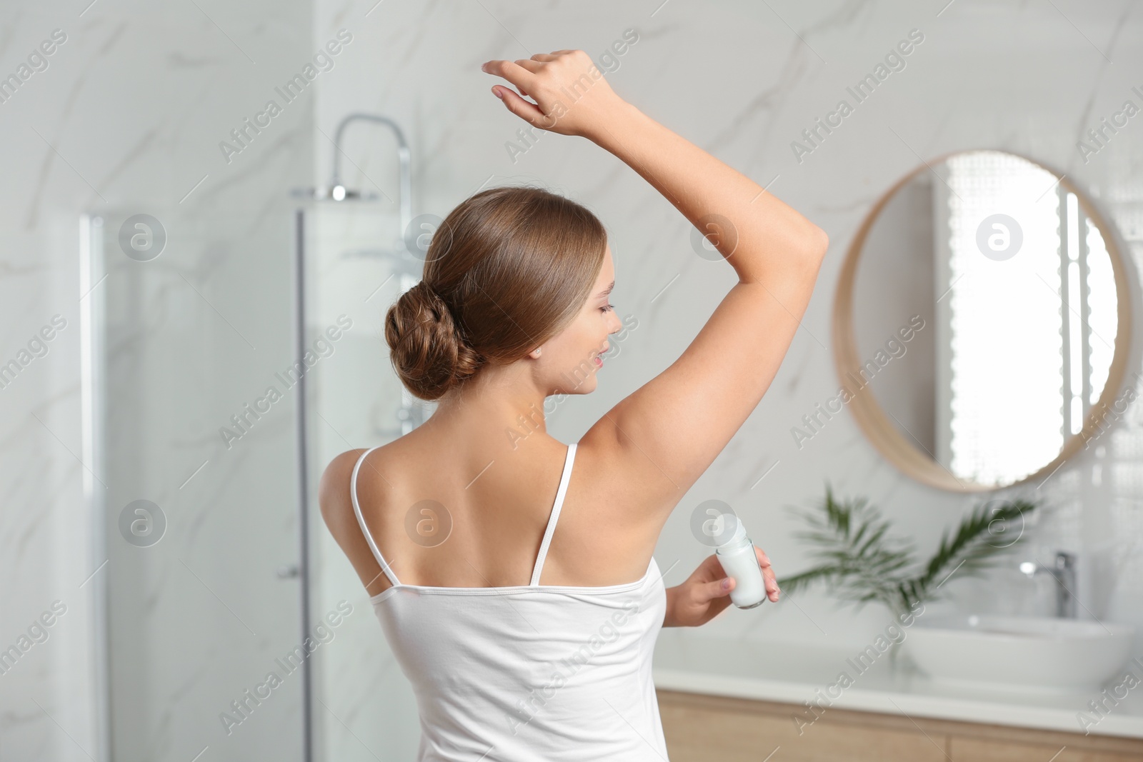 Photo of Young woman applying fresh roll-on deodorant to armpit in bathroom