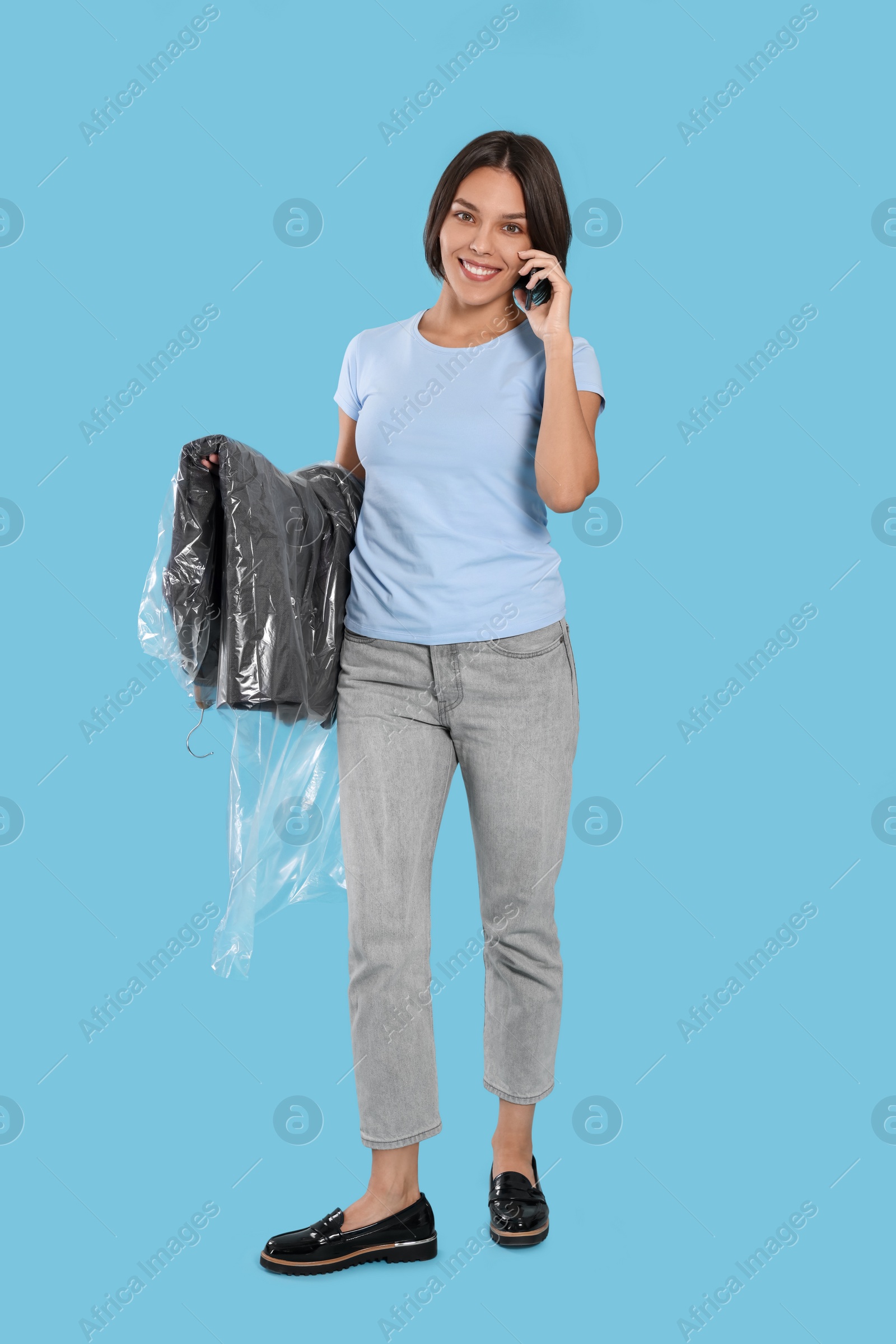 Photo of Woman holding garment cover with clothes while talking on phone against light blue background. Dry-cleaning service