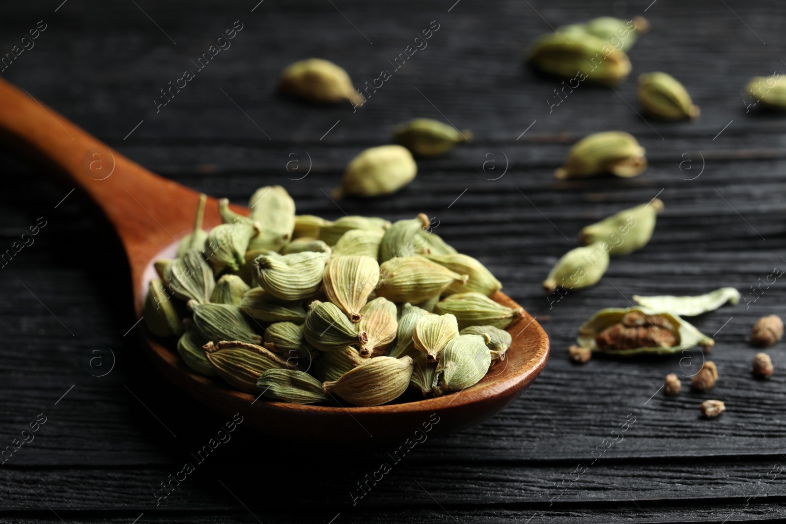 Photo of Spoon with dry cardamom pods on black wooden table, closeup