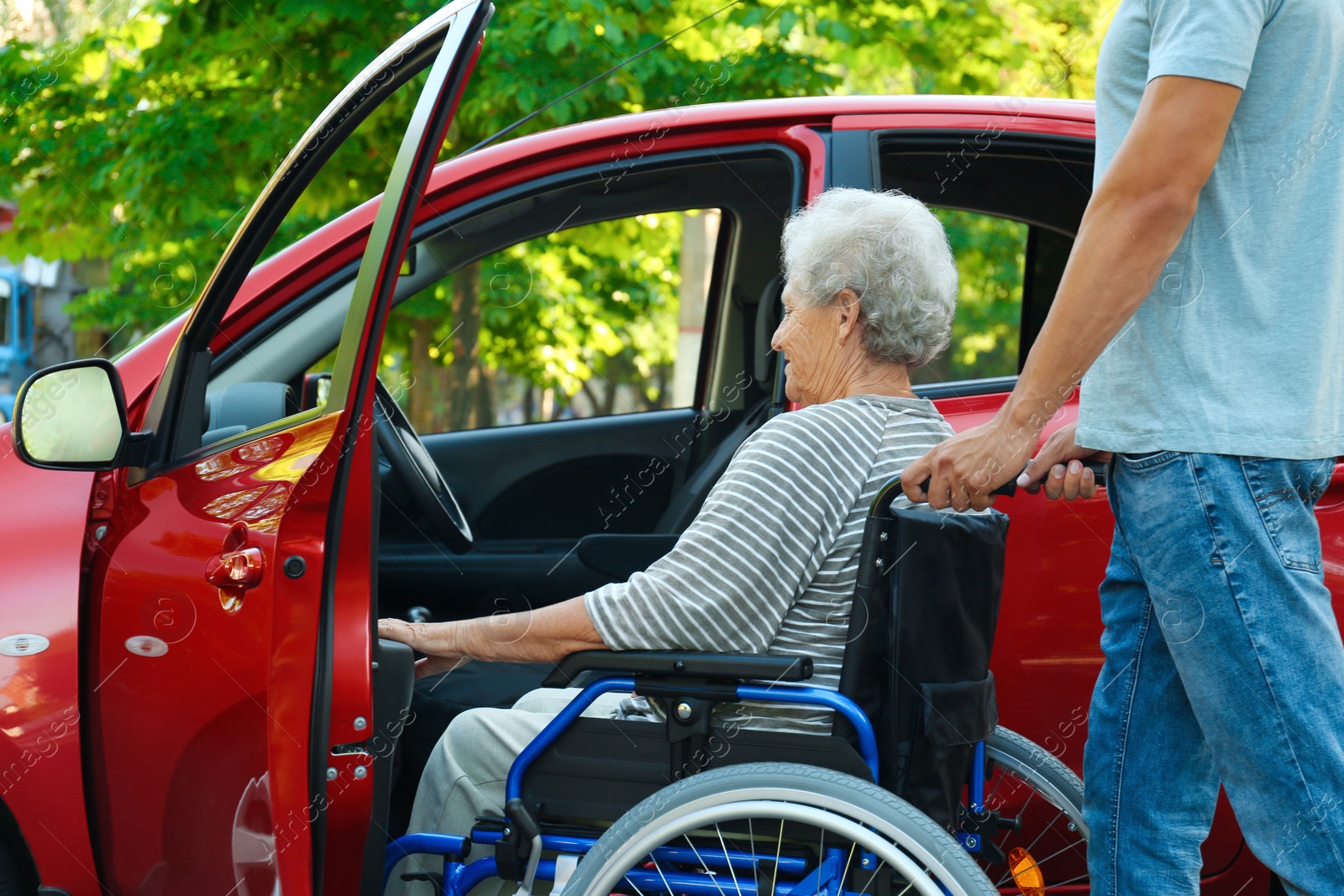 Photo of Young man helping disabled senior woman in wheelchair to get into car outdoors