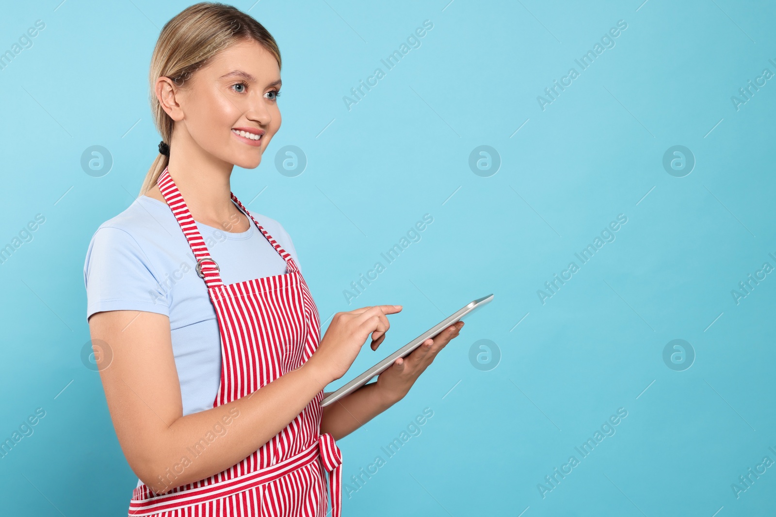 Photo of Beautiful young woman in clean striped apron with tablet on light blue background. Space for text