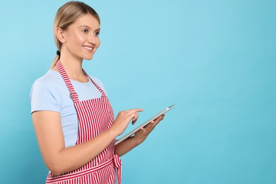 Beautiful young woman in clean striped apron with tablet on light blue background. Space for text