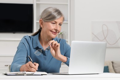 Photo of Beautiful senior woman taking notes while working with laptop at white table indoors