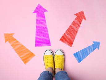 Choosing future profession. Girl standing in front of drawn signs on pink background, top view. Arrows pointing in different directions symbolizing diversity of opportunities