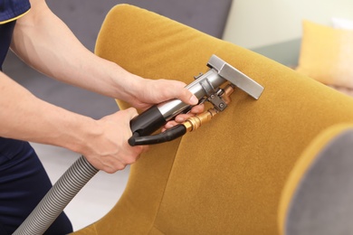Male worker removing dirt from armchair with professional vacuum cleaner indoors, closeup