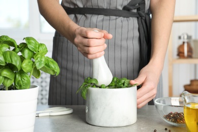 Photo of Woman grinding green basil in mortar at kitchen table