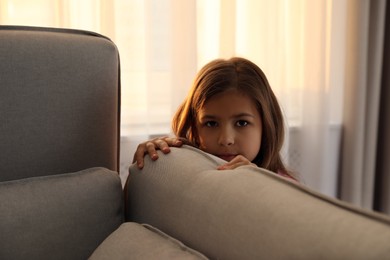 Sad little girl hiding behind sofa at home. Domestic violence concept