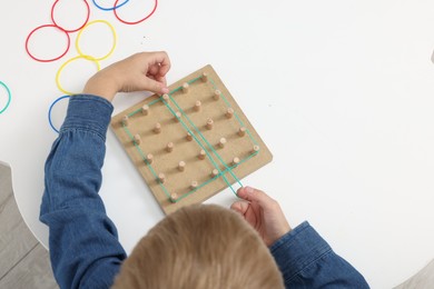 Photo of Motor skills development. Boy playing with geoboard and rubber bands at white table, top view