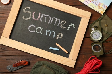 Photo of Chalkboard with text SUMMER CAMP and camping equipment on wooden background, flat lay