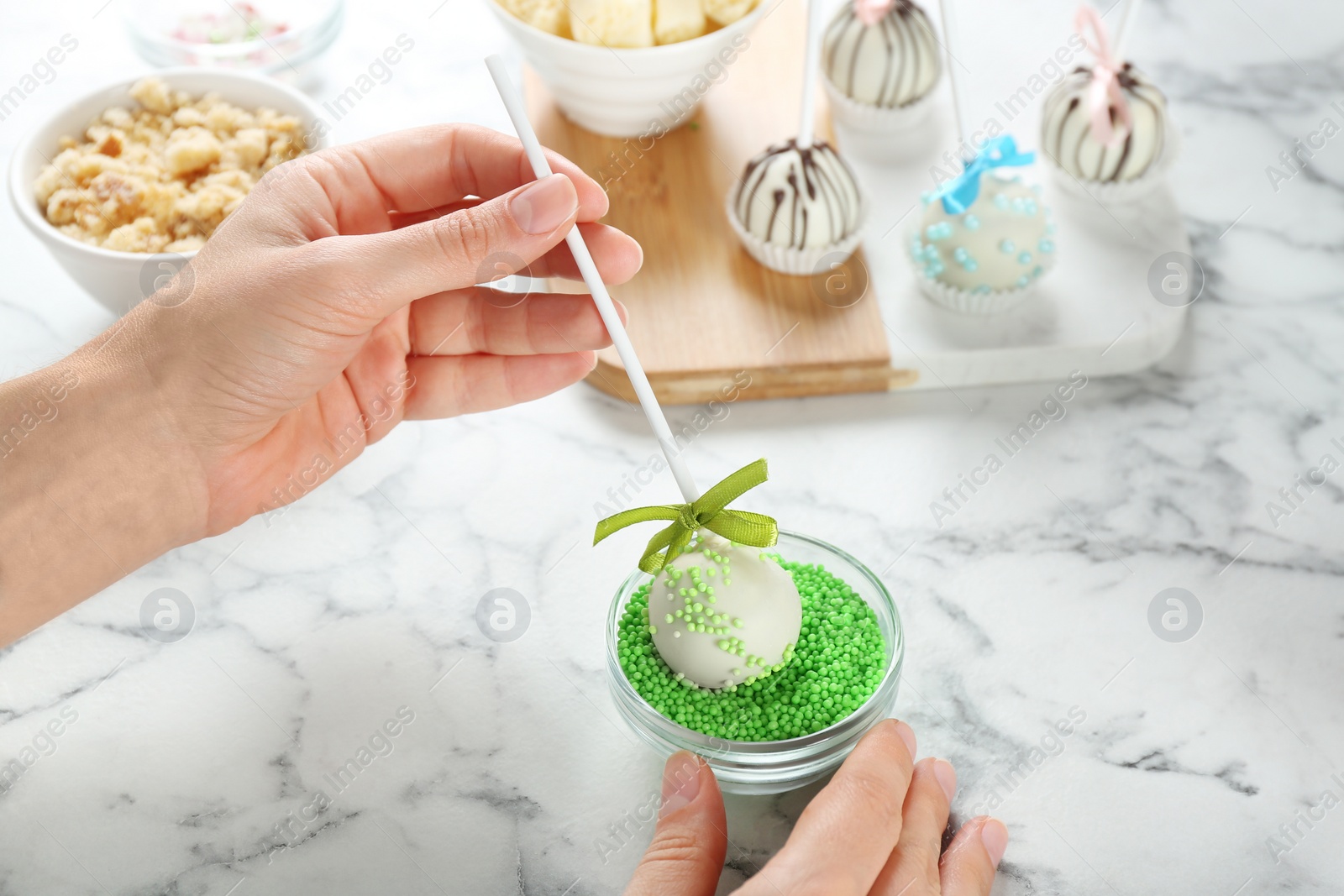 Photo of Young woman putting cake pop into green sprinkles at white marble table, closeup