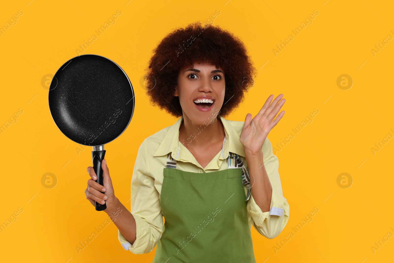 Photo of Emotional young woman in apron holding frying pan on orange background