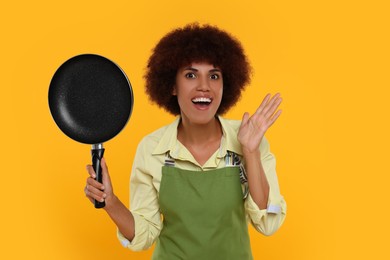 Emotional young woman in apron holding frying pan on orange background