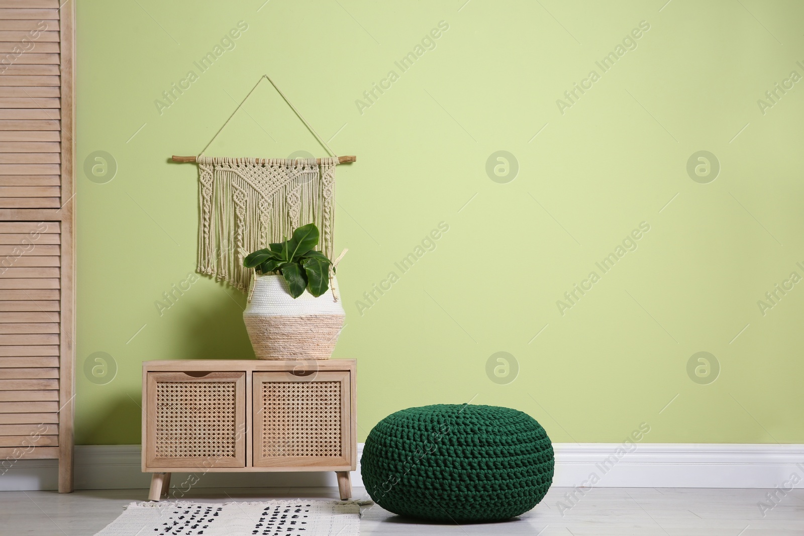 Photo of Stylish room interior with comfortable knitted pouf, wooden furniture and plant near light green wall, space for text