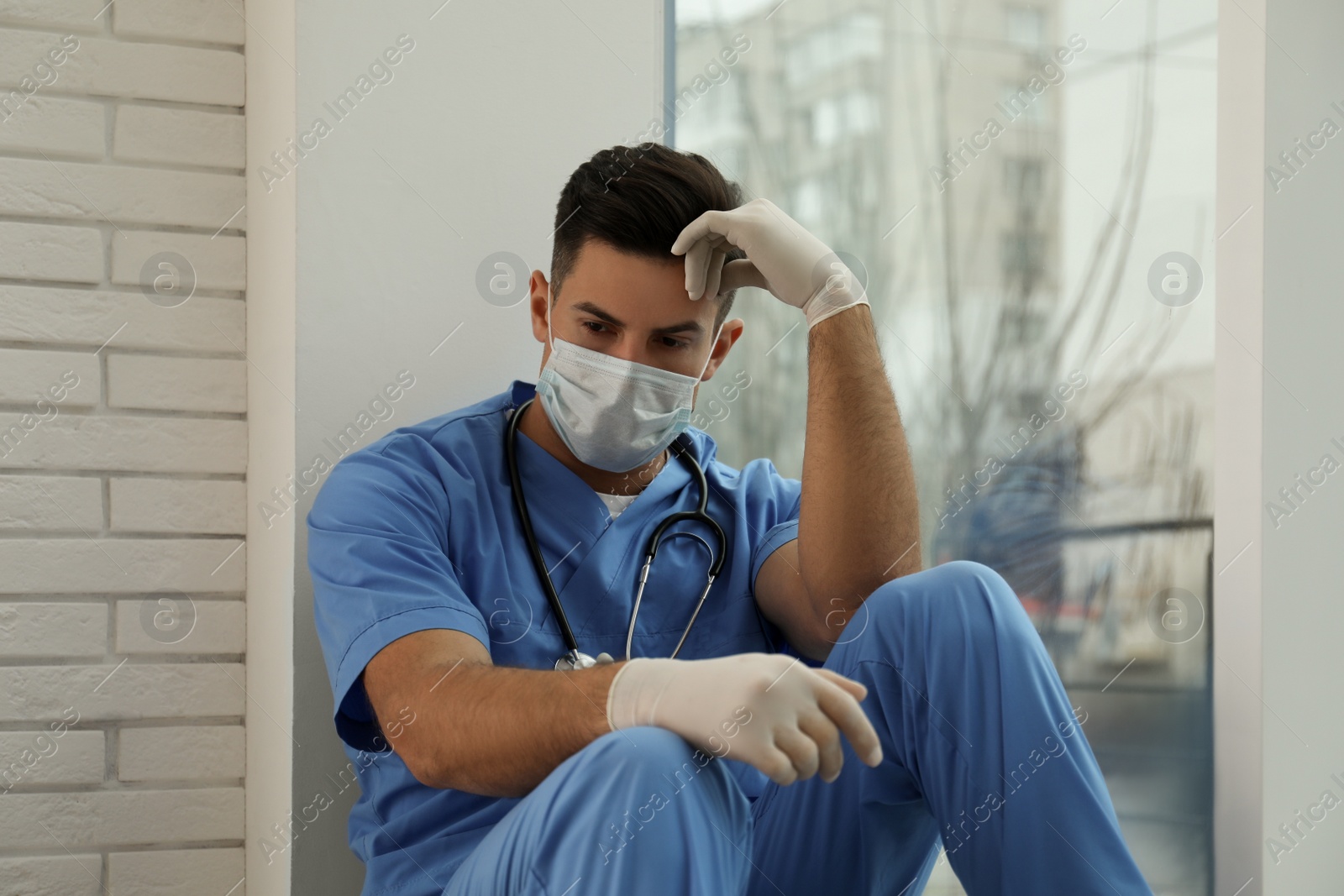 Photo of Sad doctor near window indoors. Stress of health care workers during coronavirus pandemic