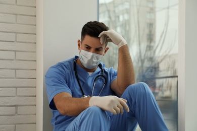 Photo of Sad doctor near window indoors. Stress of health care workers during coronavirus pandemic