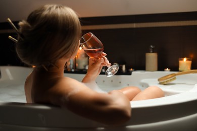 Photo of Woman drinking wine while taking bubble bath indoors, back view. Romantic atmosphere