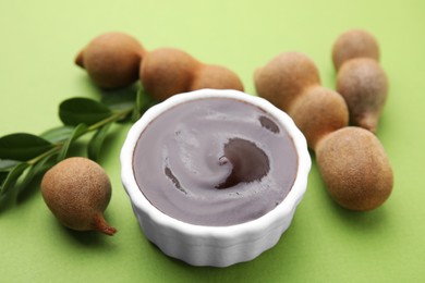 Photo of Bowl of tasty tamarind sauce, leaves and fresh pods on light green table, closeup