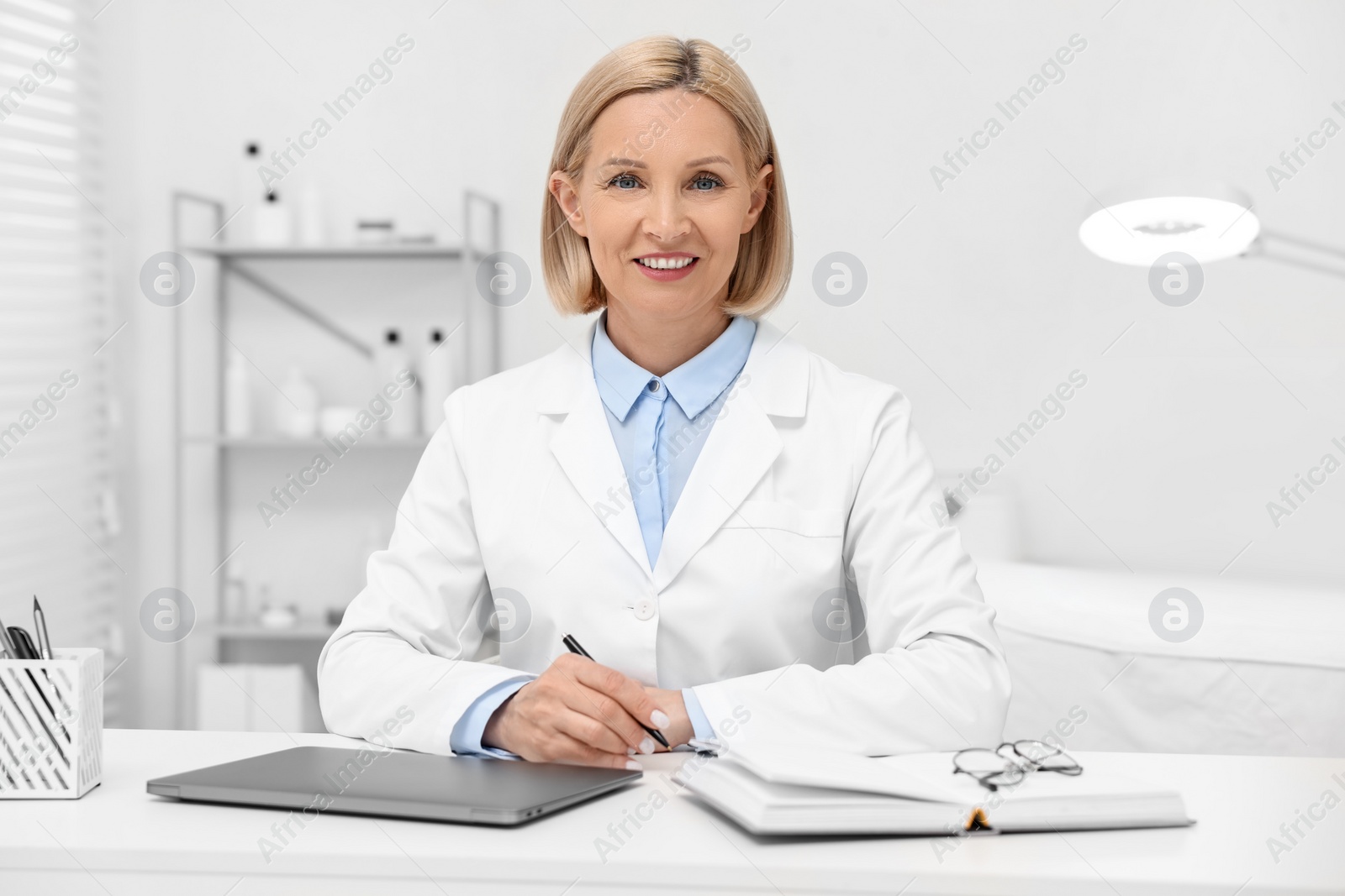 Photo of Portrait of happy dermatologist with pen at white table in clinic