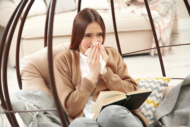 Photo of Sick young woman sneezing while reading book at home. Influenza virus