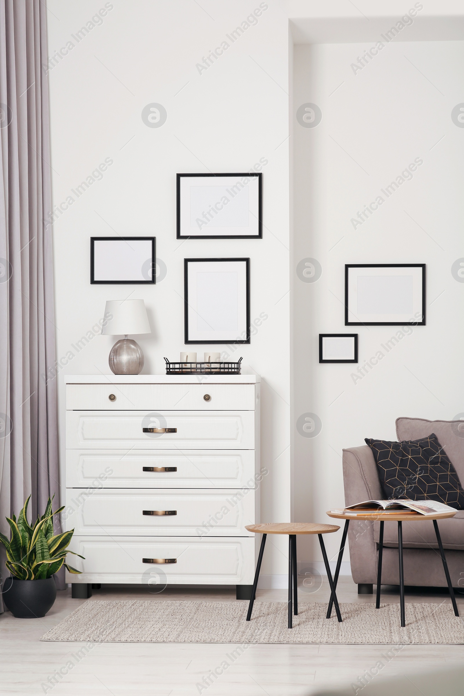 Photo of Empty frames hanging on white wall, chest of drawers and wooden tables indoors