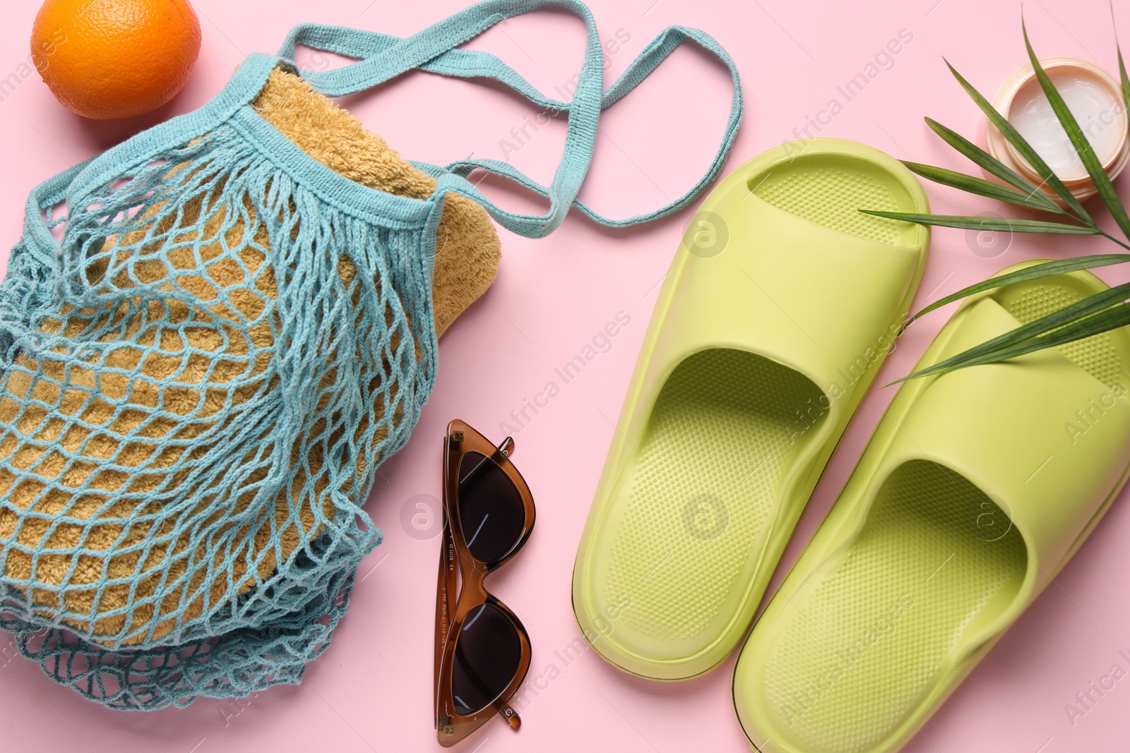 Photo of String bag with towel, sunglasses, orange, slippers, cream and palm leaf on pink background, flat lay