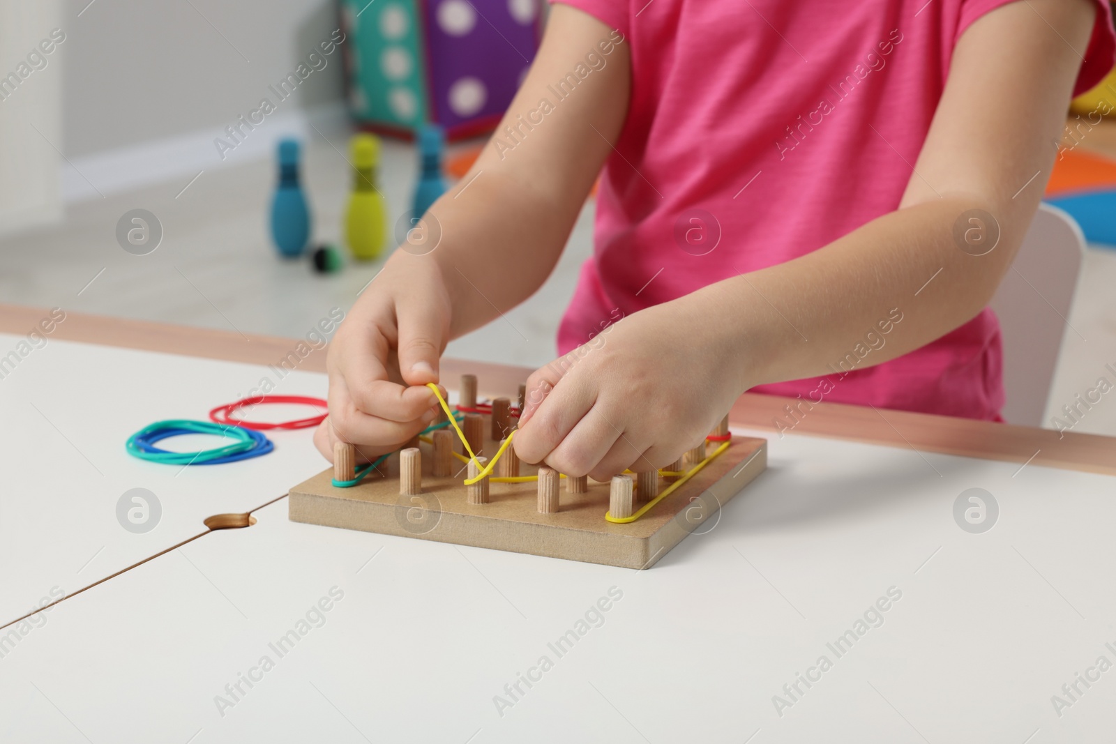 Photo of Motor skills development. Girl playing with geoboard and rubber bands at white table indoors, closeup