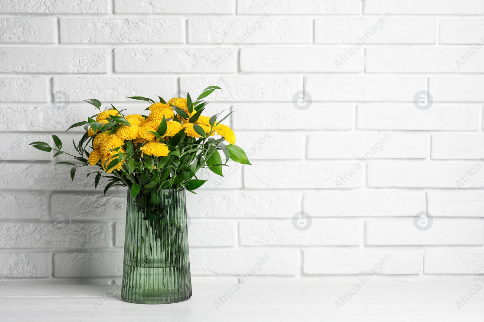 Photo of Bouquet of beautiful yellow flowers in glass vase on white wooden table near brick wall. Space for text