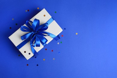 Photo of Beautiful gift box with bow and confetti on blue background, top view. Space for text
