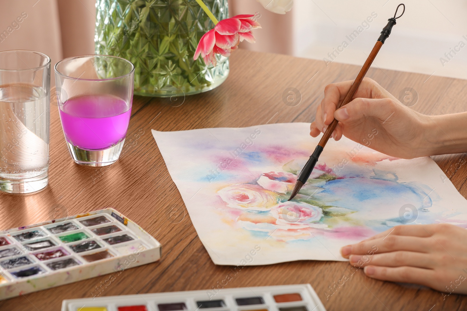 Photo of Woman painting flowers with watercolor at wooden table indoors, closeup. Creative artwork