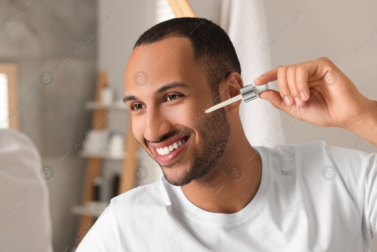 Photo of Handsome man applying cosmetic serum onto face in bathroom