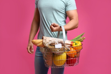 Young man with shopping basket full of products on pink background, closeup