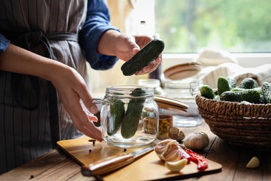 Photo of Woman putting cucumbers into jar at wooden table, closeup. Pickling vegetables