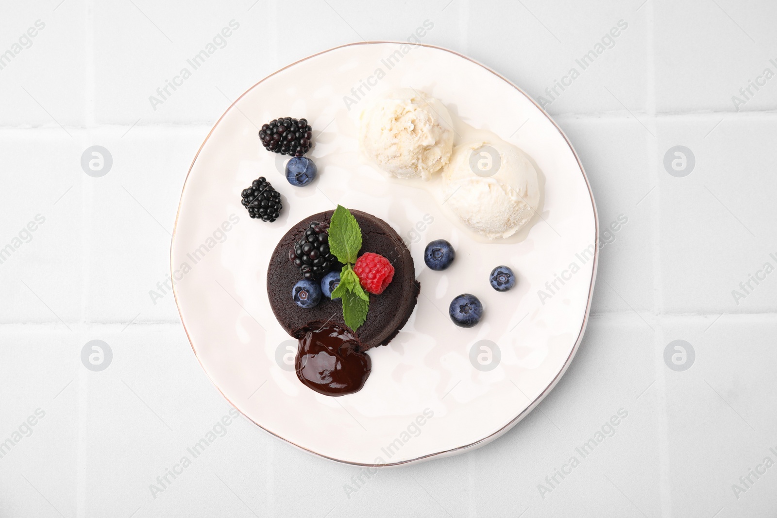 Photo of Plate with delicious chocolate fondant, berries, ice cream and mint on white tiled table, top view