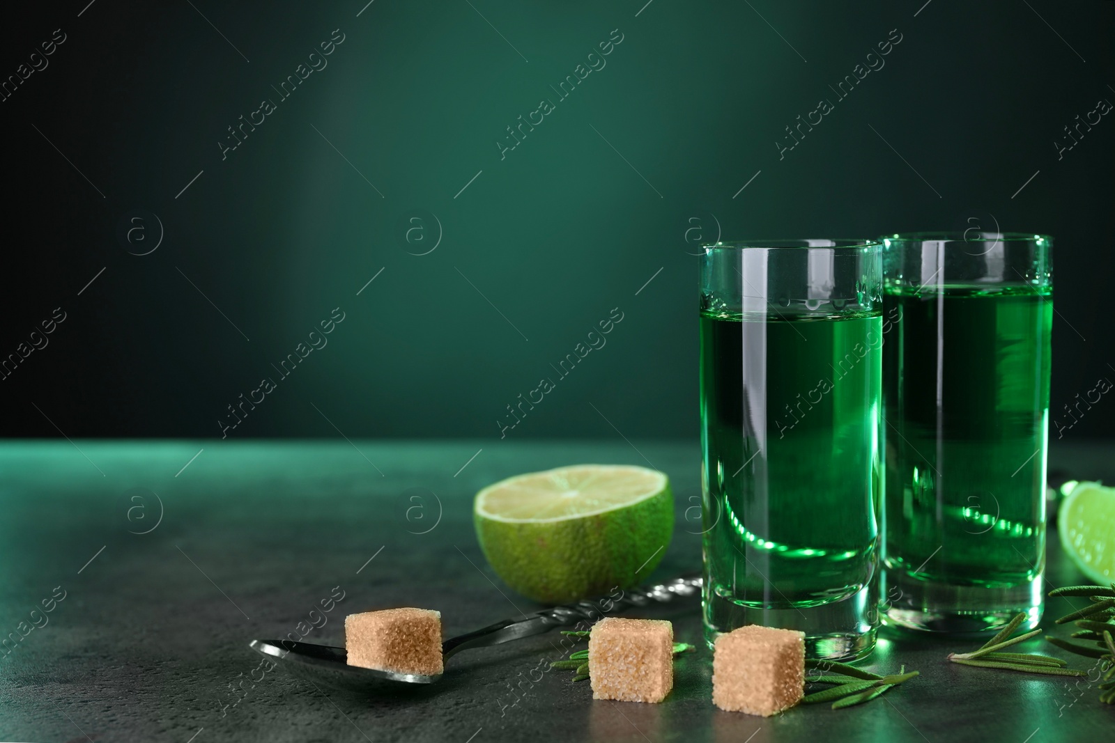 Photo of Absinthe in shot glasses, spoon, brown sugar, lime and rosemary on gray textured table against green background, space for text. Alcoholic drink