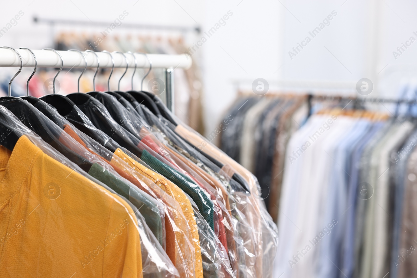 Photo of Dry-cleaning service. Many different clothes in plastic bags hanging on rack indoors, closeup