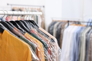 Photo of Dry-cleaning service. Many different clothes in plastic bags hanging on rack indoors, closeup