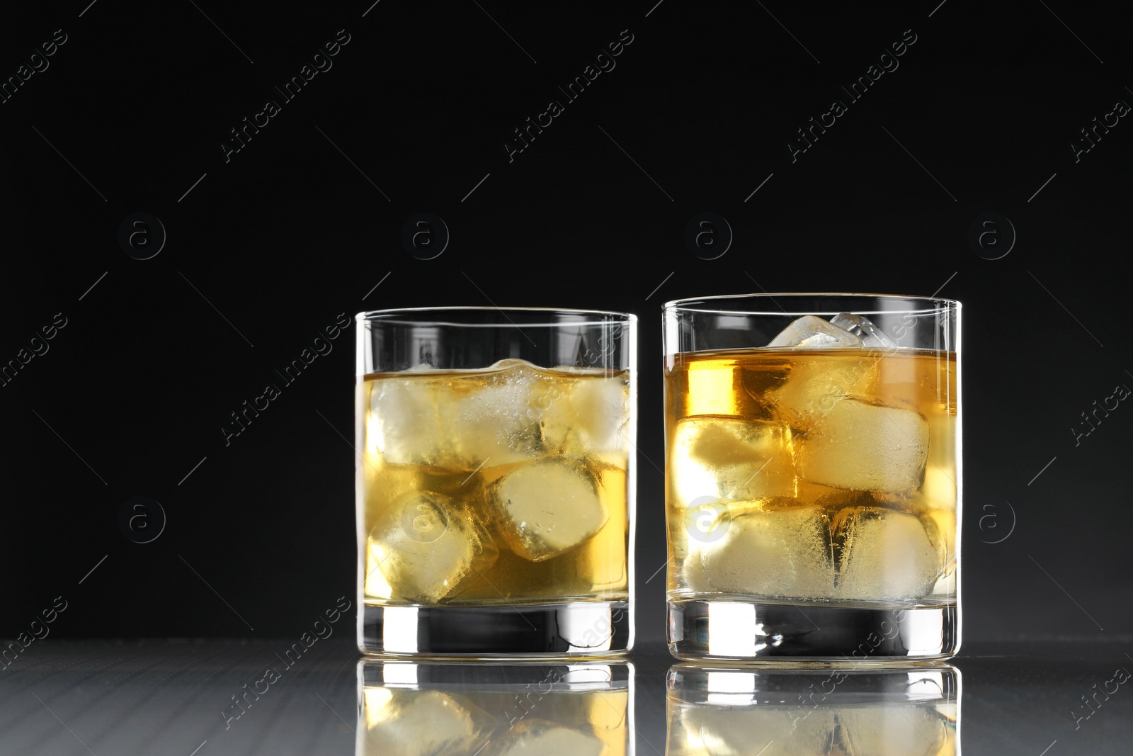 Photo of Tasty whiskey with ice in glasses on mirror table against black background, closeup