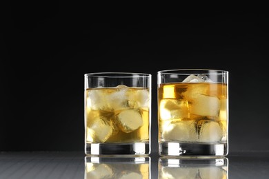 Tasty whiskey with ice in glasses on mirror table against black background, closeup