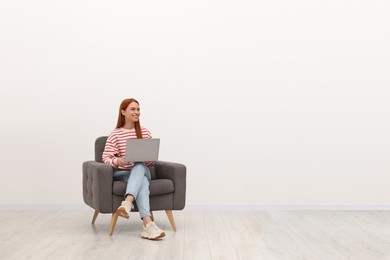 Photo of Happy young woman with laptop sitting in armchair near white wall indoors. Space for text