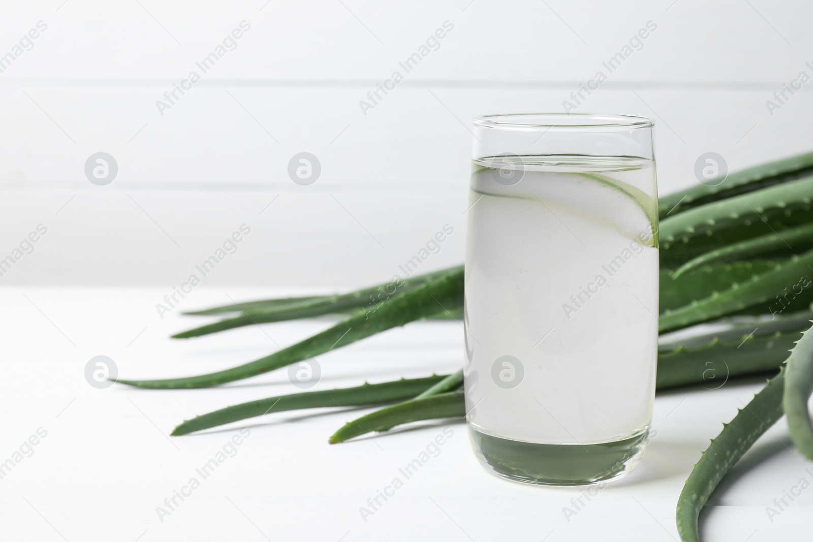 Photo of Fresh aloe juice in glass and leaves on white wooden table. Space for text