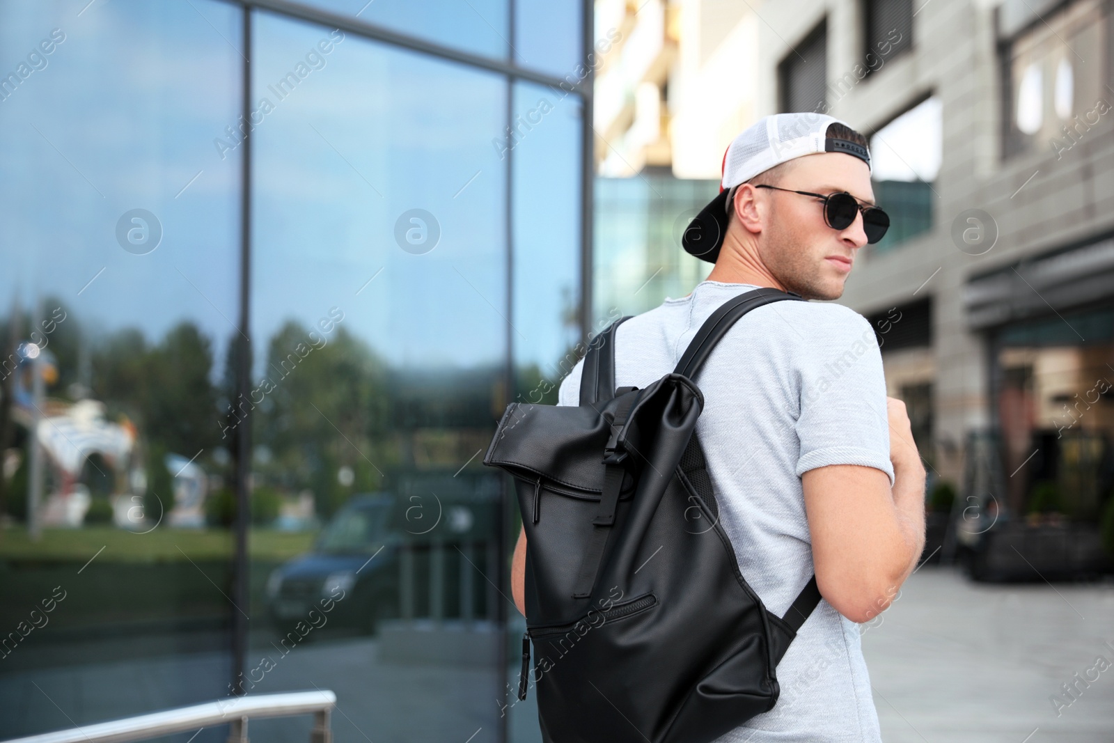 Photo of Handsome young man with stylish sunglasses and backpack near building outdoors, space for text