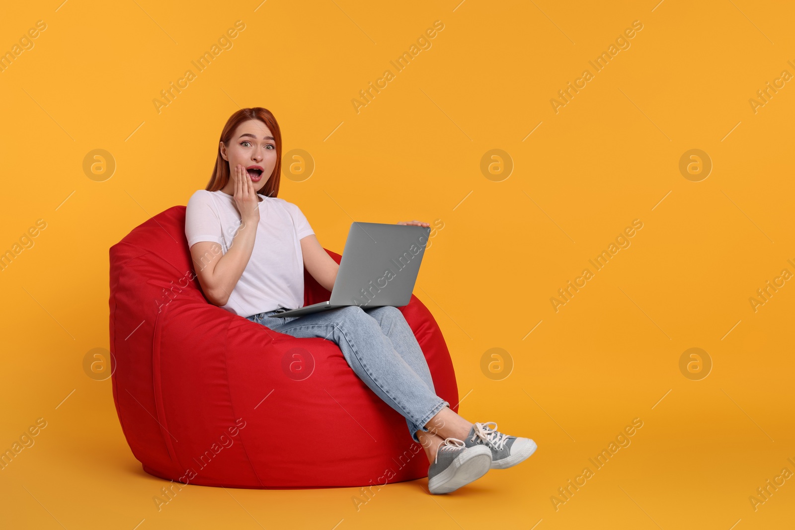 Photo of Surprised young woman with laptop sitting on beanbag chair against yellow background