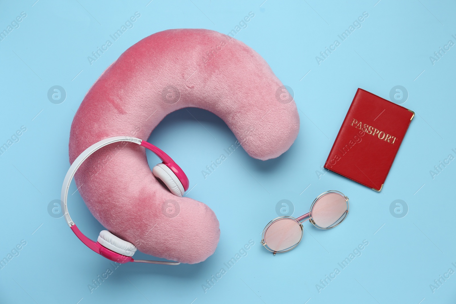 Photo of Pink travel pillow with headphones, passport and sunglasses on light blue background, flat lay