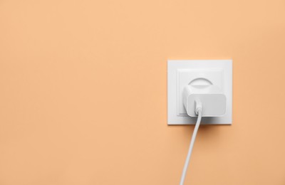 Photo of Charger adapter plugged into power socket on pale orange wall, space for text. Electrical supply
