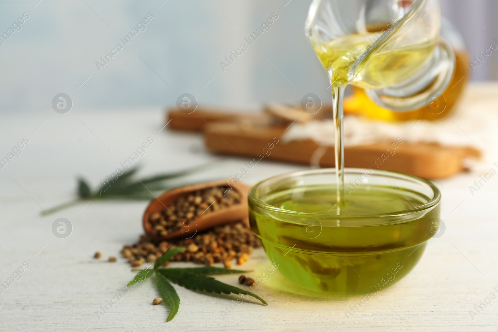 Photo of Hemp oil pouring from sauce boat into bowl on white table