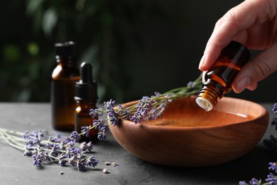 Photo of Woman dripping essential oil from bottle into bowl near lavender at grey table, closeup. Space for text