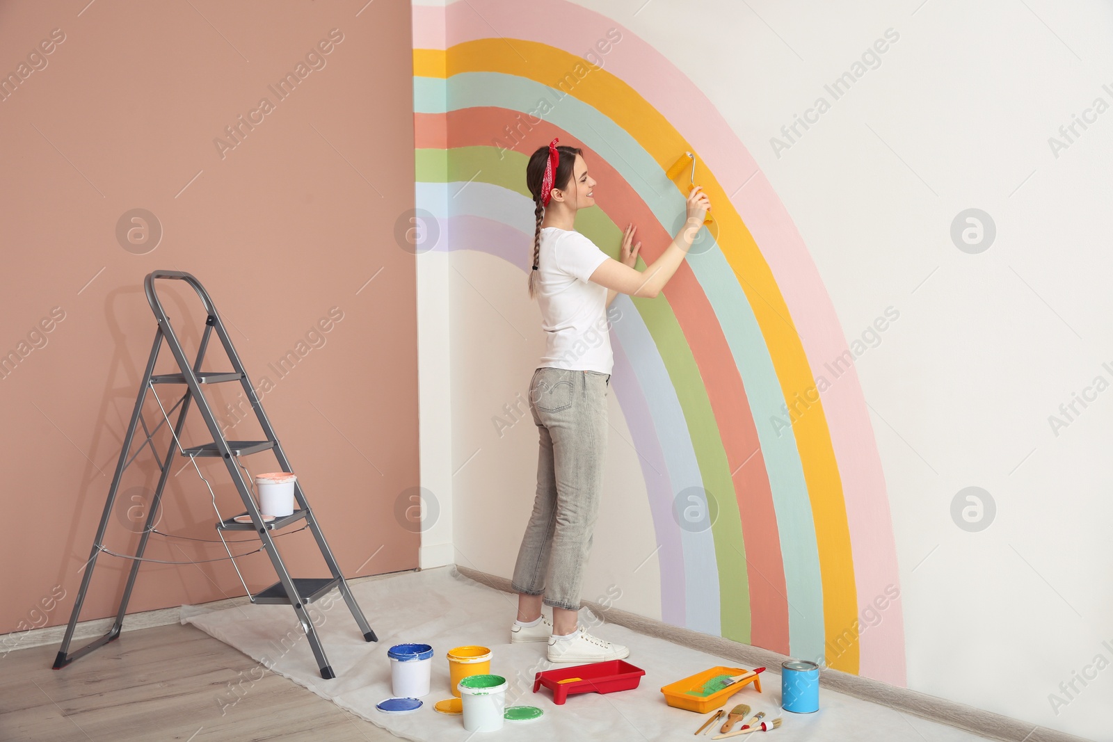 Photo of Young woman painting rainbow on white wall indoors