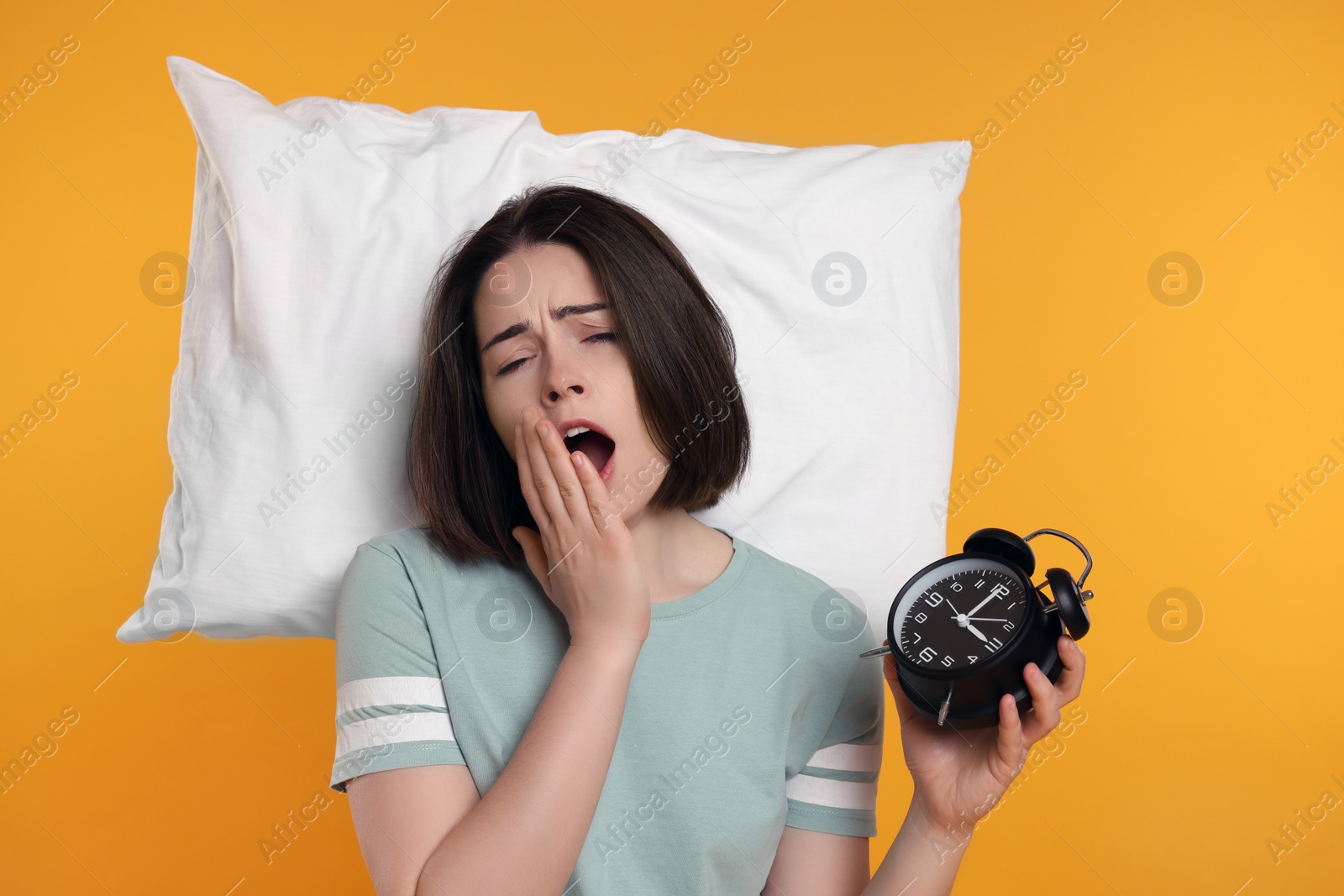 Photo of Sleepy young woman with alarm clock and pillow yawning on yellow background. Insomnia problem